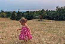 Photo Pink Dress by Lydia Miller. A photo of a young girl with short blond hair and a bright pink dress in a field of tall grass with a blue sky.