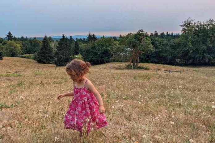 Photo Pink Dress by Lydia Miller. A photo of a young girl with short blond hair and a bright pink dress in a field of tall grass with a blue sky.