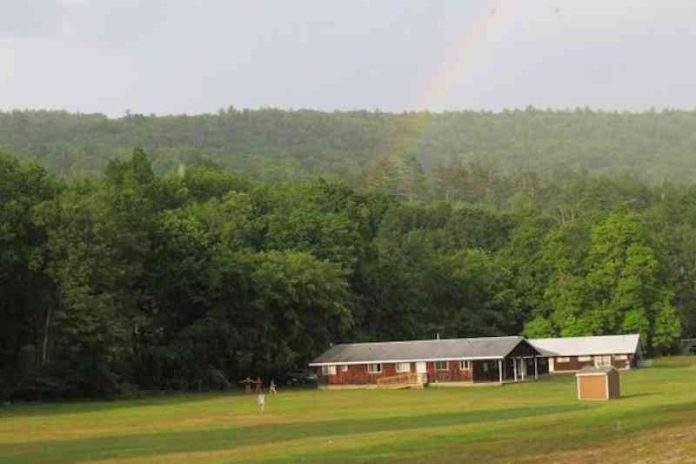 A photo of a person walking down a grassy hill a small one-story house in front of a forest.