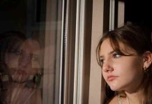 Teenage girl stares thoughtfully at her reflection in the window which shows a pondering and somewhat sad expression.