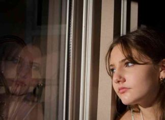 Teenage girl stares thoughtfully at her reflection in the window which shows a pondering and somewhat sad expression.