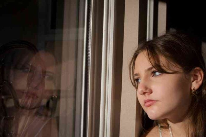 Teenage girl stares thoughtfully at her reflection in the window which shows a pondering and somewhat sad expression.