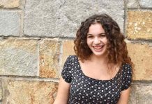 A girl with curly brown hair smiles at the camera. She is standing in front of a stone wall.