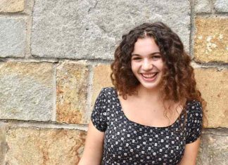 A girl with curly brown hair smiles at the camera. She is standing in front of a stone wall.