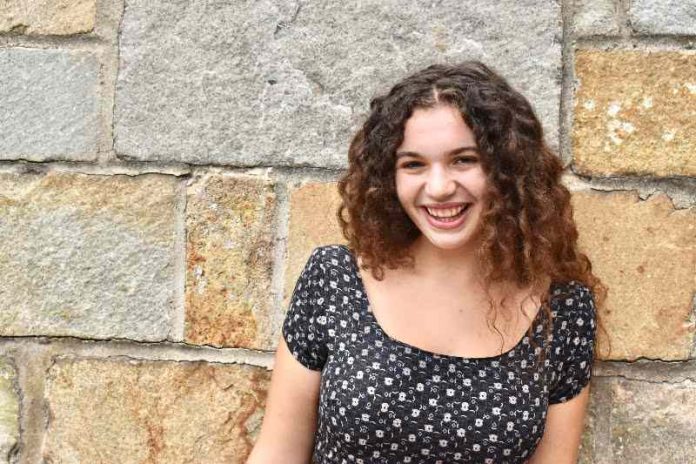 A girl with curly brown hair smiles at the camera. She is standing in front of a stone wall.