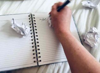 A hand scribbles in a notebook surrounded by crumpled-up pieces of paper.