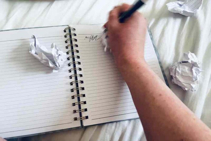 A hand scribbles in a notebook surrounded by crumpled-up pieces of paper.