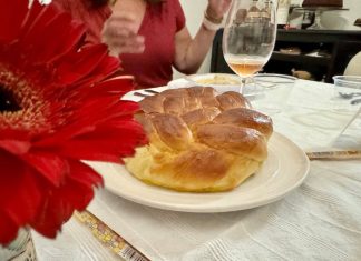 A round-braided challah loaf, on a table surrounded by wine glasses, cutlery, and a red flower.