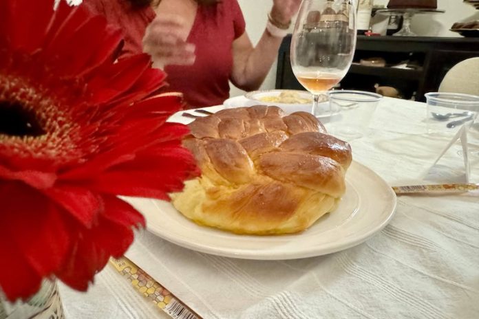 A round-braided challah loaf, on a table surrounded by wine glasses, cutlery, and a red flower.