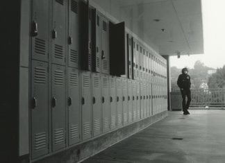A boy dressed in a black hoodie stands on an outdoor balcony in front of a row of open lockers.