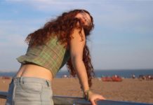 A girl with curly red hair leans back on a fence with the beach behind her.