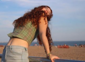 A girl with curly red hair leans back on a fence with the beach behind her.
