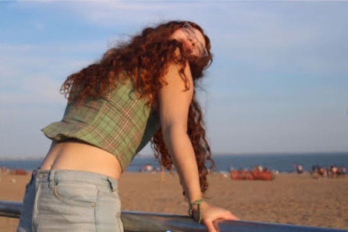 A girl with curly red hair leans back on a fence with the beach behind her.