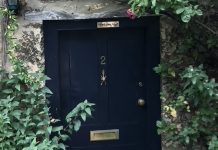 An angled shot of a black door set into a concrete wall that is framed by flowers. The door is numbered with a golden 2 and includes a letter box and door knocker shaped like an insect. A plaque at the top reads "Mind your head."