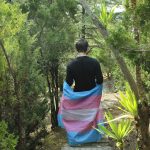 A person with short brown hair walks along a hiking trail, their back to the camera. They are wearing a black long-sleeve shirt with a trans pride flag wrapped around their waist.