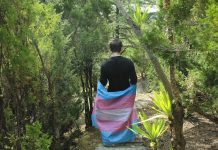 A person with short brown hair walks along a hiking trail, their back to the camera. They are wearing a black long-sleeve shirt with a trans pride flag wrapped around their waist.