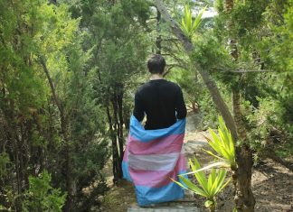 A person with short brown hair walks along a hiking trail, their back to the camera. They are wearing a black long-sleeve shirt with a trans pride flag wrapped around their waist.