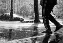 A black and white photo of a person's legs, walking down a sidewalk through heavy rain.