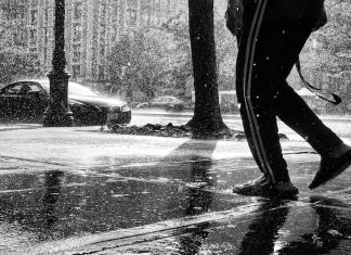 A black and white photo of a person's legs, walking down a sidewalk through heavy rain.