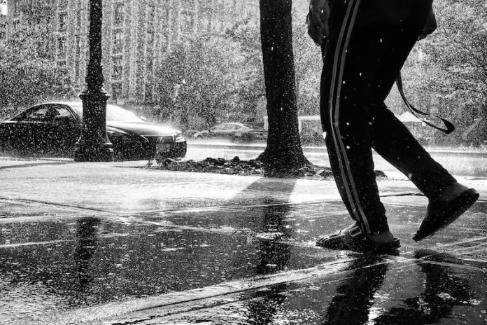 A black and white photo of a person's legs, walking down a sidewalk through heavy rain.