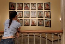 A girl leans on a railing at the top of a wide, curved stairwell. Her back is to the camera, and she’s looking at a gallery wall of portraits of Indigenous Americans on the landing beneath her.