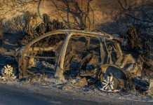 All that remains of a sedan is its metal frame, which is sinking into a pile of debris at the side of the road. The rusted color of the frame matches the rust-red dirt and bushes that surround it.