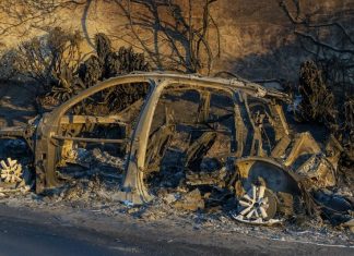 All that remains of a sedan is its metal frame, which is sinking into a pile of debris at the side of the road. The rusted color of the frame matches the rust-red dirt and bushes that surround it.