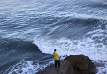 A man in a yellow raincoat stands on a cliff edge, overlooking crashing waves.