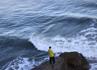 A man in a yellow raincoat stands on a cliff edge, overlooking crashing waves.