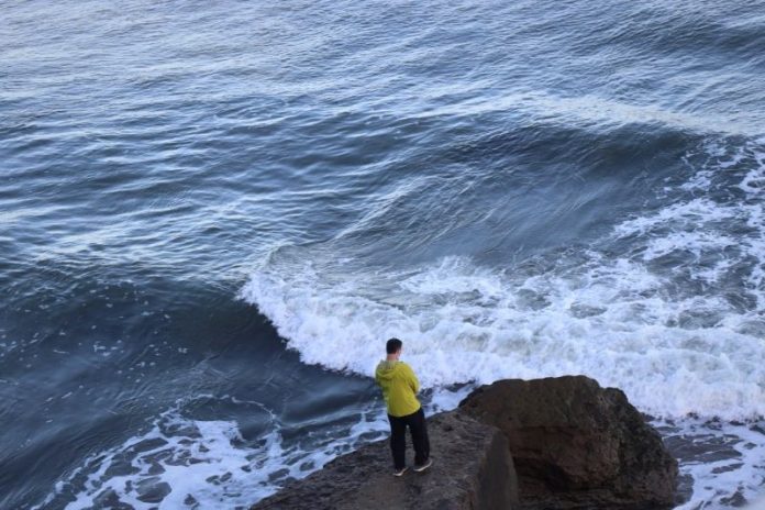 A man in a yellow raincoat stands on a cliff edge, overlooking crashing waves.
