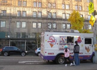 A young girl wearing a helmet and an older person holding a scooter talk to an ice cream truck driver on a city street in front of a large scaffolded building.
