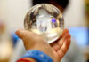 A hand holding a clear glass sphere, with a blurred background and a person’s face partially visible through the sphere.