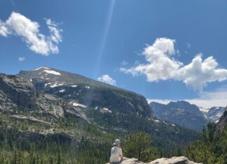 A teen boy sitting on a boulder admiring the mountains, trees, and blue sky in the valley around him