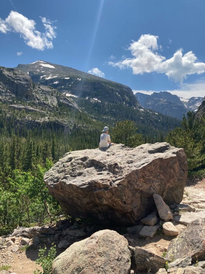 A teen boy sitting on a boulder admiring the mountains, trees, and blue sky in the valley around him