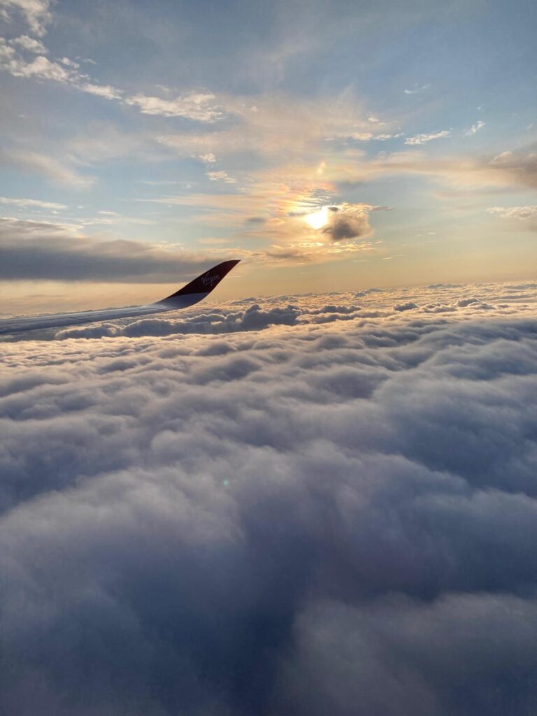 The wing of a plane flies through a sea of puffy clouds, with the setting sun giving the whole image a peachy glow