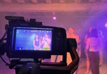 Directed By? A camera films a group of people in a dimly lit room with purple lighting; the camera display shows two women posing.