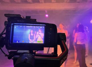 A camera films a group of people in a dimly lit room with purple lighting; the camera display shows two women posing.