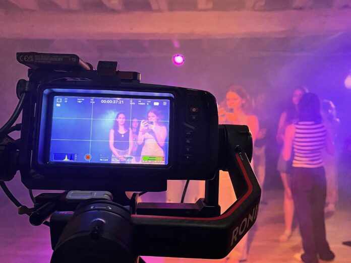 A camera films a group of people in a dimly lit room with purple lighting; the camera display shows two women posing.