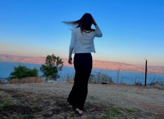 A girl, viewed from behind, looks out over a vibrant blue lake at sunset. Her long brown hair floats in the wind.