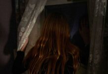 A shiny-haired girl looks out a window with her hand pressed on it.