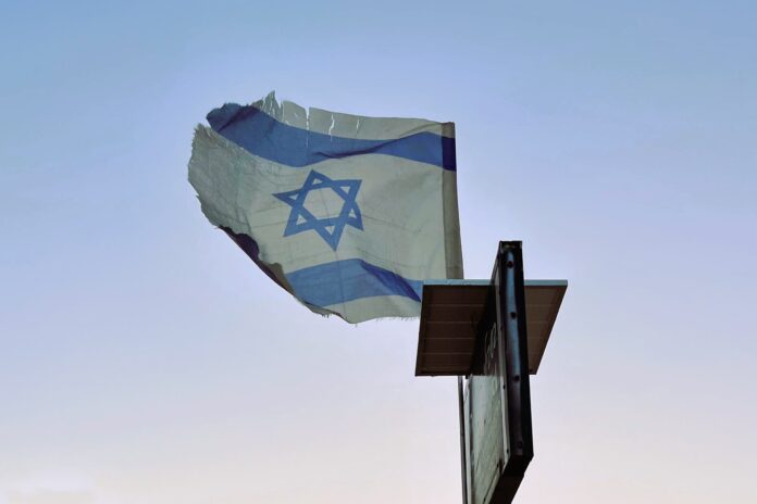 A tattered Israeli flag flies on a post against a pale blue sky.
