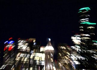 A blurry scene of a city at night. Buildings glow in the darkness.