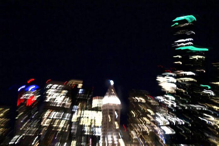 A blurry scene of a city at night. Buildings glow in the darkness.