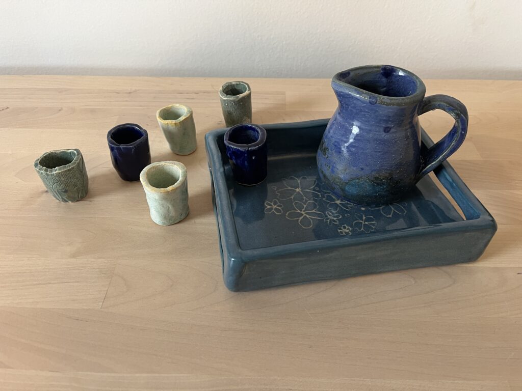 Naama Raun's Kiddush set includes a blue pitcher with five small individual cups. The pitcher and one of the individual cups sits on a ceramic tray decorated with floral print.