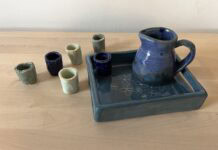 Naama Raun's Kiddush set includes a blue pitcher with five small individual cups. The pitcher and one of the individual cups sits on a ceramic tray decorated with floral print.