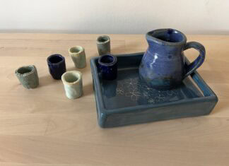 Naama Raun's Kiddush set includes a blue pitcher with five small individual cups. The pitcher and one of the individual cups sits on a ceramic tray decorated with floral print.