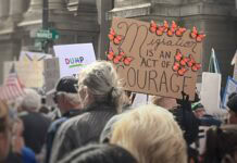 Protesters march near a cardboard sign that says "Migration is an act of courage" surrounded by monarch butterflies.