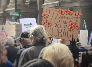 Protesters march near a cardboard sign that says "Migration is an act of courage" surrounded by monarch butterflies.