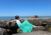 A young woman in a turquoise skirt lounges on a rocky outcrop beside a body of water. A lone seagull flies overhead.