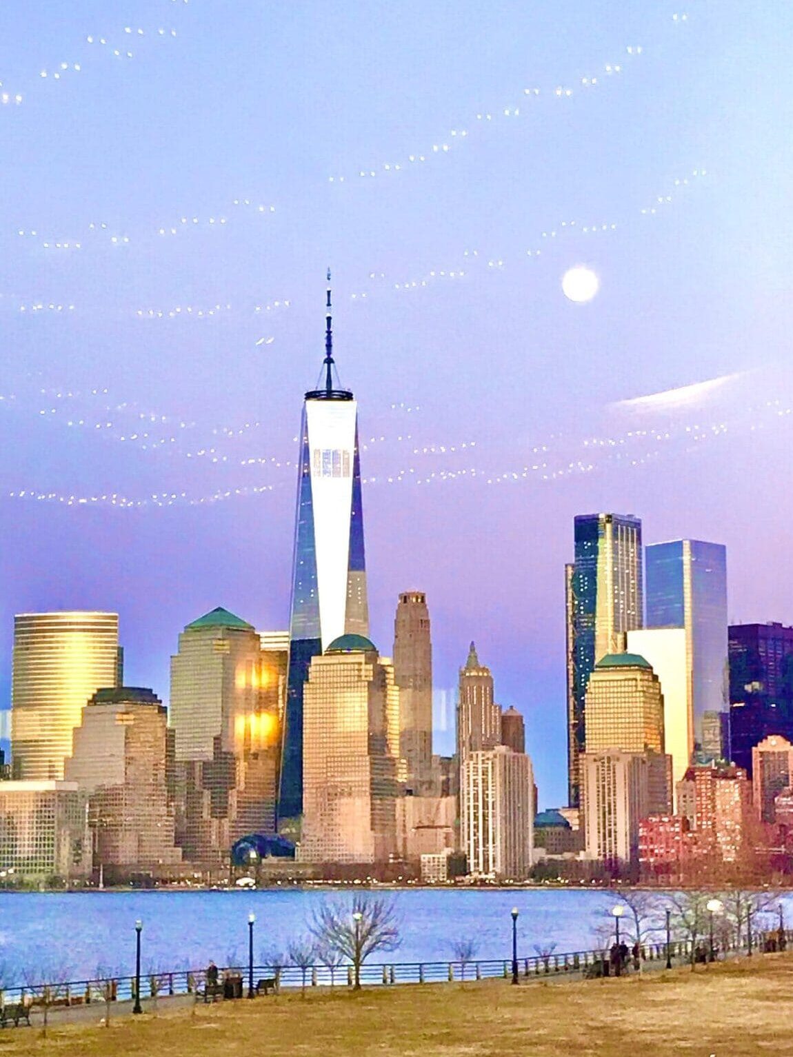 The sun sets in the purple and blue sky behind tall buildings in Liberty State Park.